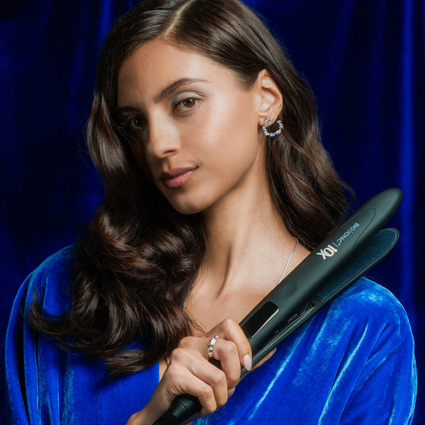 Model with wavy hair holding the 10X Styling Iron on a blue velvet background